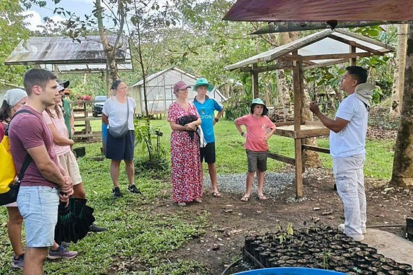 A group of people standing outdoors near a wooden structure with a roof, surrounded by greenery and a dirt ground. 2