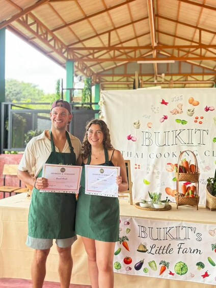 Two people holding certificates in front of a banner with the text 'BUKIT THAI COOKING CLUB' and 'BUKIT'S Little Farm'. 2