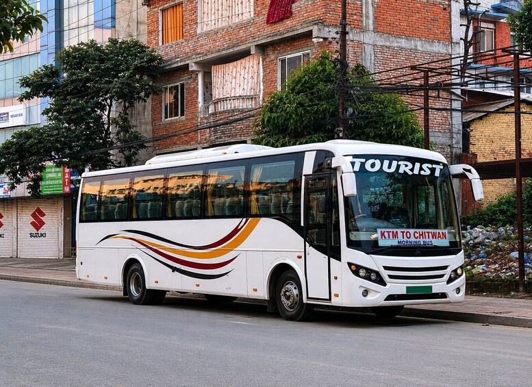 A white bus with 'TOURIST' and 'KTMTO CHITWAN MORNING BUS' on its front, parked on a street with buildings in the background. 1
