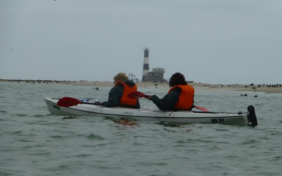 Two people in a white canoe with orange life vests on a calm body of water, a lighthouse in the background, and a sandy beach with people on the shore. 2