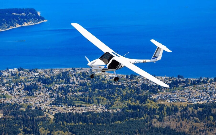 A small white aircraft flying over a coastal town with dense forest and a clear blue sea. 3