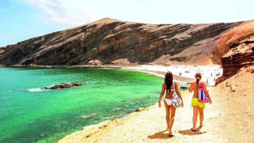 Two people walking along a beach with clear turquoise water and rocky cliffs in the background. 1