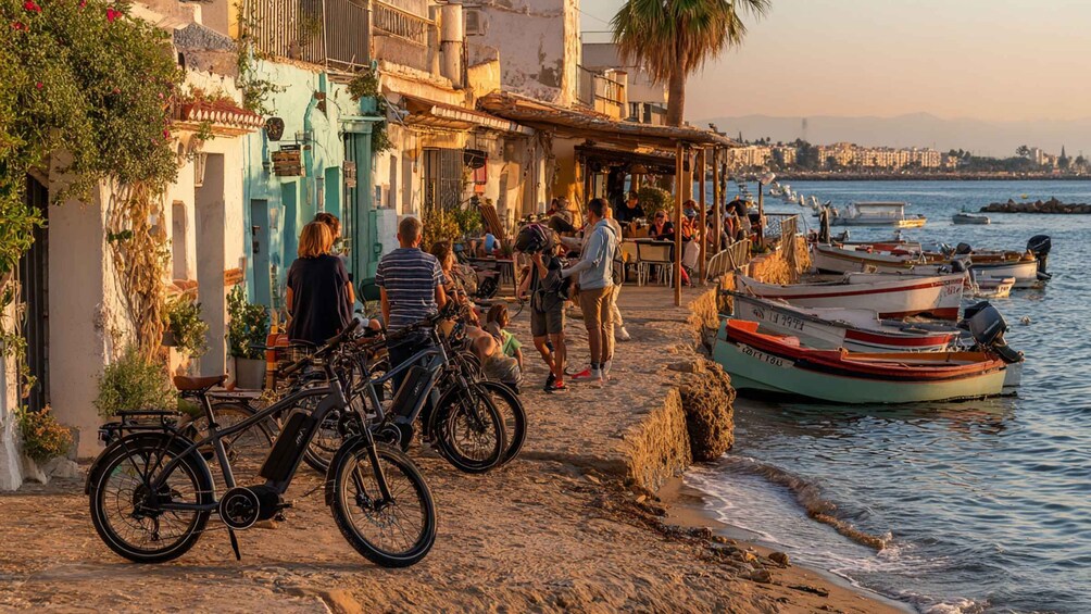A coastal town with people walking along a stone path, bicycles parked, and boats in the water. 5