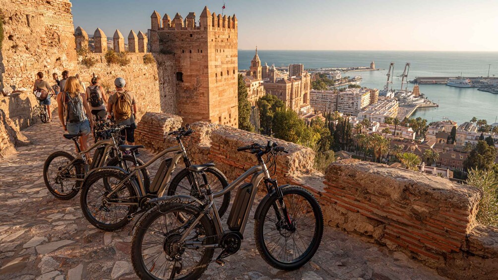 A historic stone wall with bicycles parked in front, overlooking a coastal city with a prominent castle and port. 3