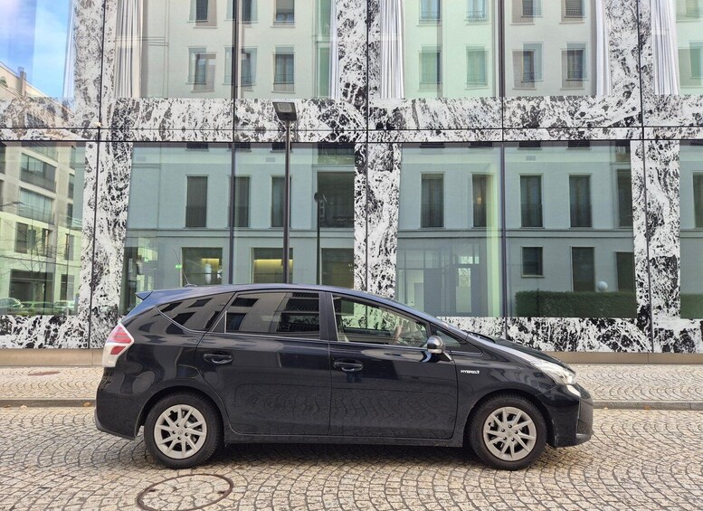 A black hybrid car parked on a cobblestone street in front of a modern building with large windows and a marble facade. 5