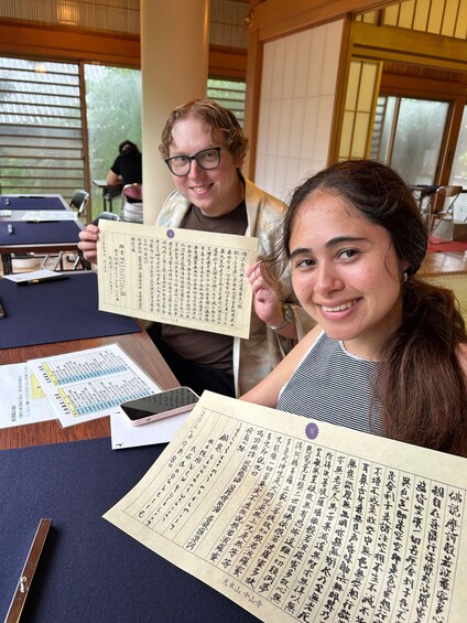 Two individuals holding large sheets of paper with Japanese text, in a room with wooden furniture and a view of greenery outside. 2