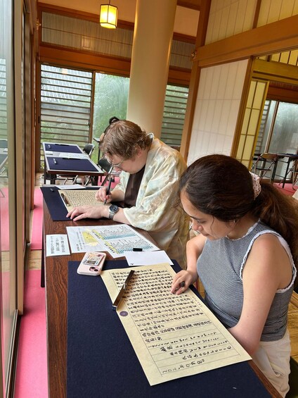 Two individuals writing on large sheets of paper at a table with traditional Japanese sliding doors in the background. 1
