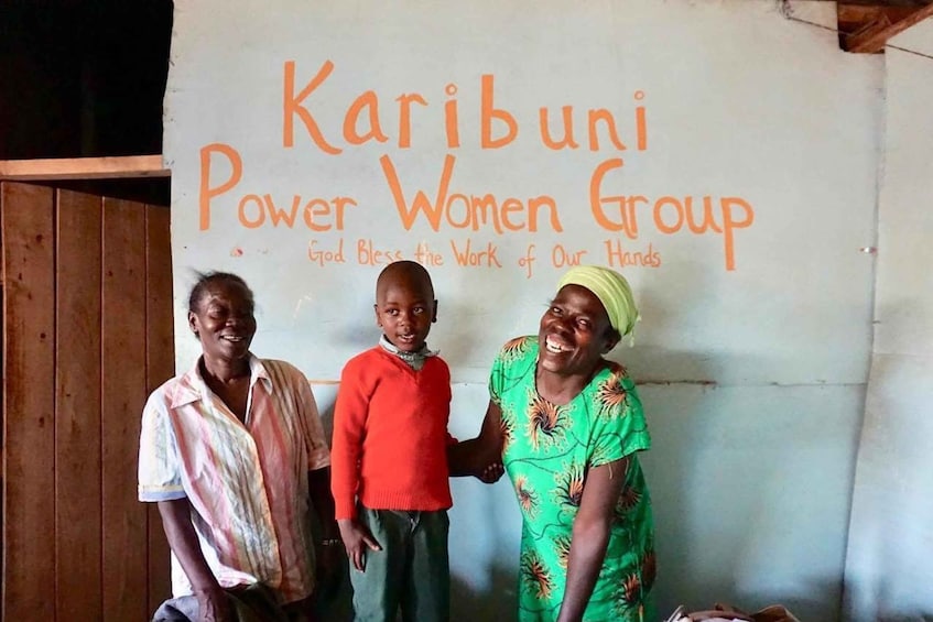Three people standing in front of a wall with the text "Karibuni Power Women Group" and a slogan. 3