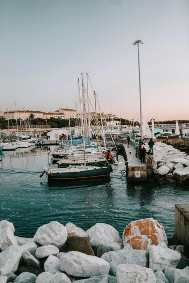A marina with boats docked, people walking, and large rocks in the foreground. 2