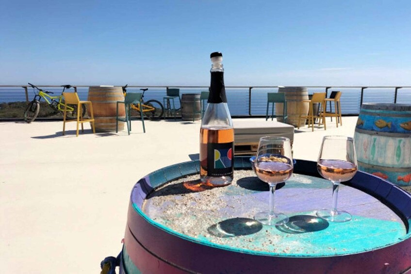 A table with a bottle and two glasses of wine, chairs, and a bicycle on a beach with the sea in the background. 1