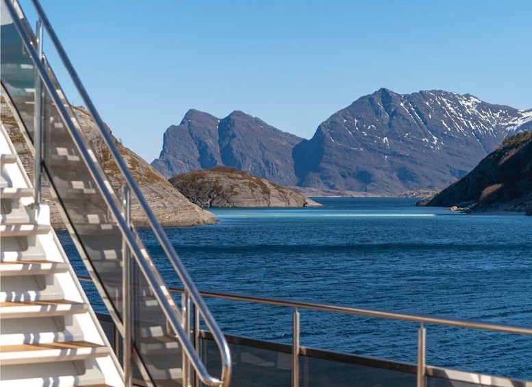 A ship's deck with metal railings overlooking a calm sea and snow-capped mountains in the background. 1