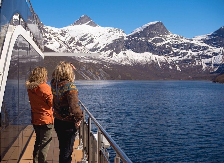 Two people on a boat deck overlooking a glacier-covered mountain range and a calm body of water. 4