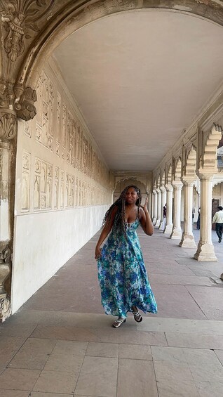 A woman in a blue floral dress walking in a corridor with ornate columns and detailed wall carvings. 4