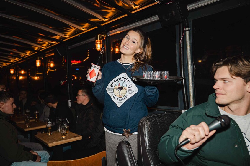 A person holding a bottle of beer and a tray with glasses, in a dimly lit indoor setting with tables and chairs. 4