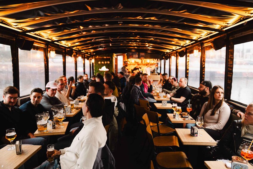 A boat interior with people seated at tables, holding drinks, and a bar area in the background. 1