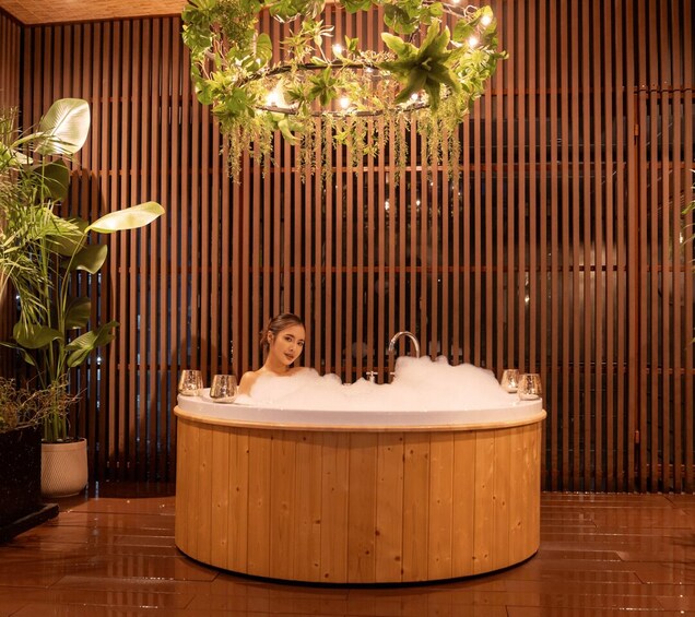 A woman in a wooden hot tub with bubbles, surrounded by plants and a wooden slat wall. 2