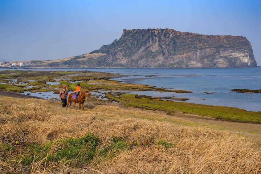 Paisaje costero con un acantilado destacado, un jinete y una persona caminando junto al caballo. 5