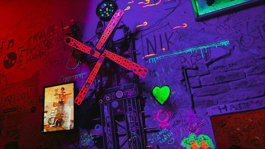 A graffiti-covered wall with a neon X, a green heart, and a poster of a drummer. 5