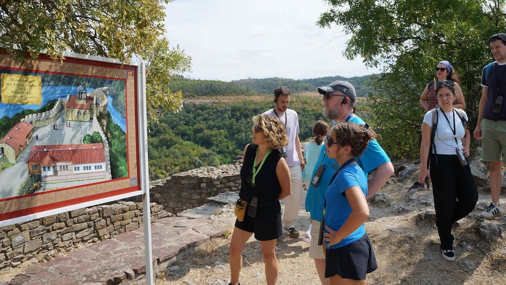 A group of people standing near a historical site with a mural depicting a castle and a river. 2