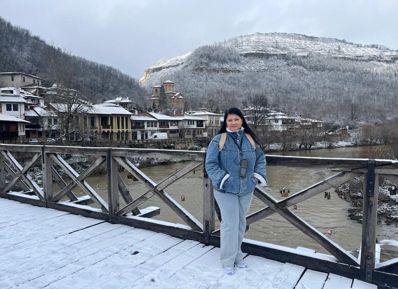 A person standing on a snow-covered wooden bridge with a river and buildings in the background. 5