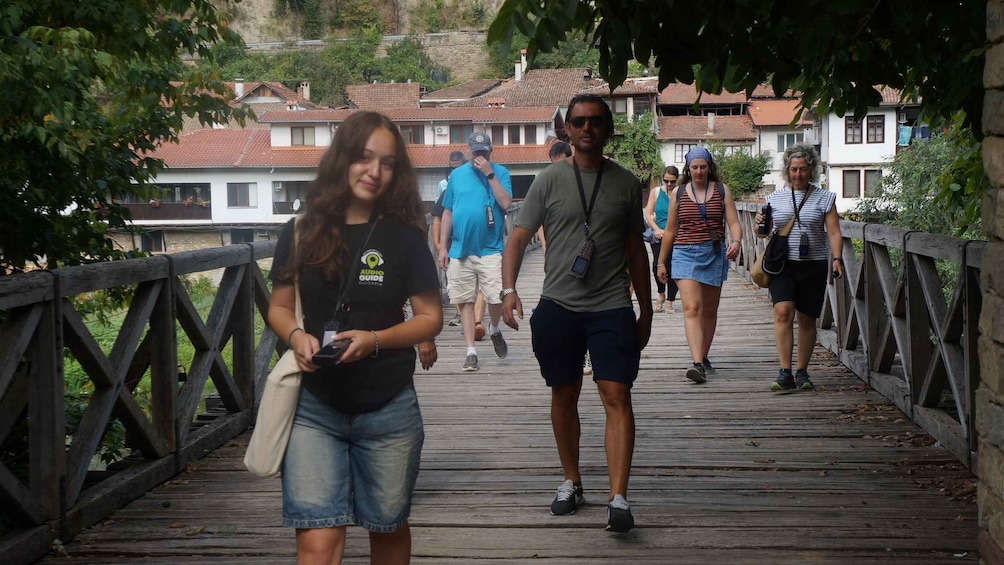 A wooden bridge with people walking on it, surrounded by greenery and buildings in the background. 3