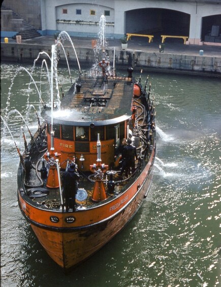 A vintage fireboat named 'Freda Busse' with water jets spraying from its deck, docked near a bridge with a white building in the background. 1