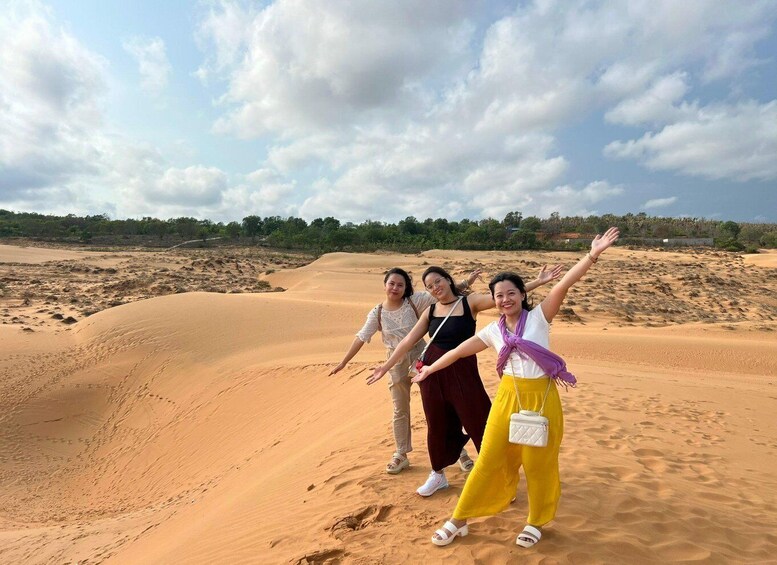 Three people standing on a sand dune with their arms raised, surrounded by a desert landscape with sparse vegetation and a partly cloudy sky. 5