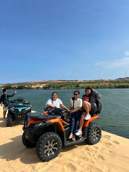 Three people on a quad bike near a lake with wind turbines in the background. 3