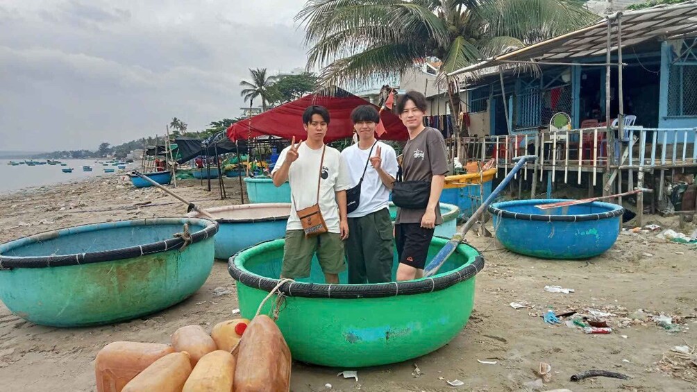 Three people standing in a green boat on a beach with several other boats and a makeshift shelter in the background. 2