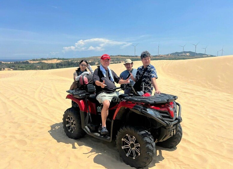 A group of people on a red ATV in a desert with sand dunes and wind turbines in the background. 1