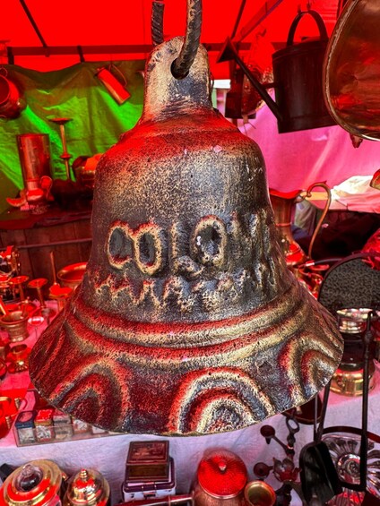 A large, ornate bell with intricate carvings and a handle, displayed in a market setting with various items in the background. 5