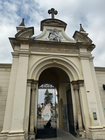 A white building with a cross on top and an archway with a black gate. 1