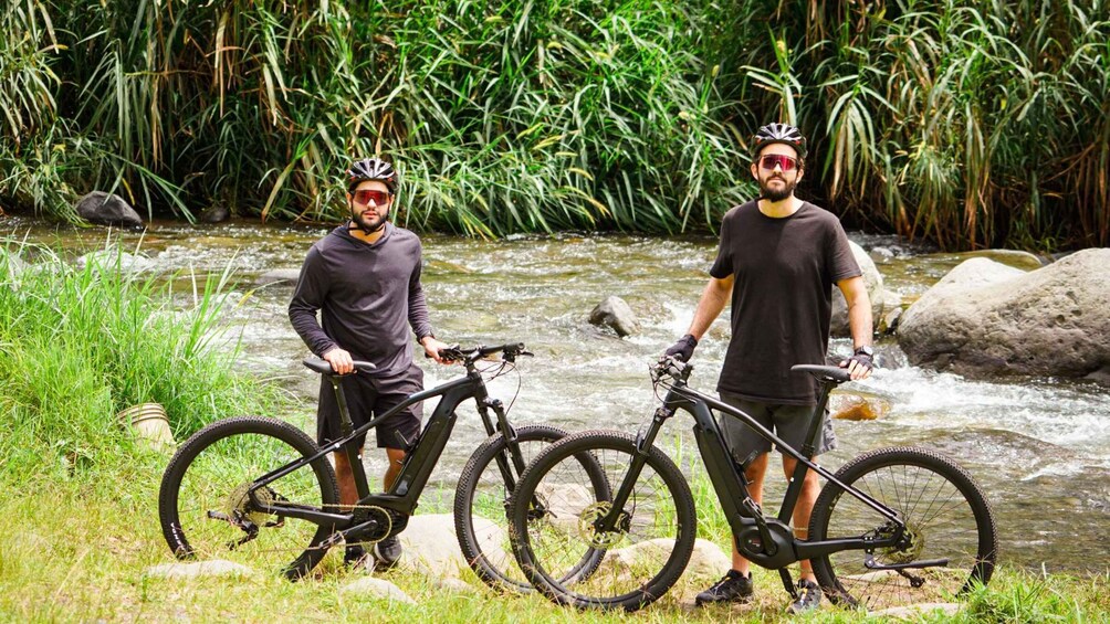Two cyclists standing by a flowing river with dense greenery in the background. 5
