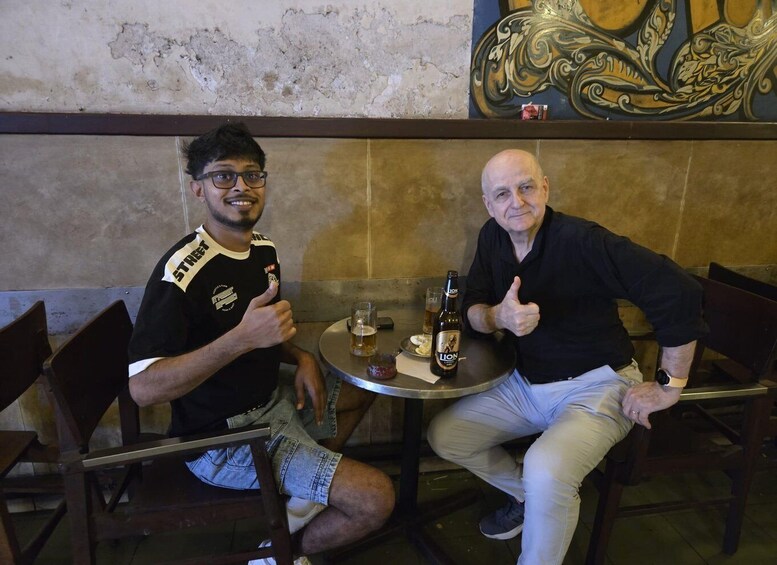 Two people sitting at a table with drinks, a decorative wall, and a chair. 2