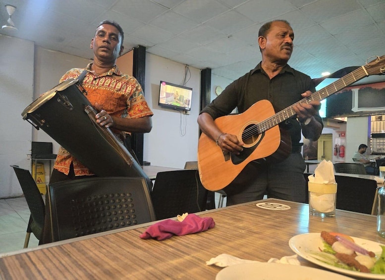 Two men playing guitars in a room with a table, chairs, and a television. 4