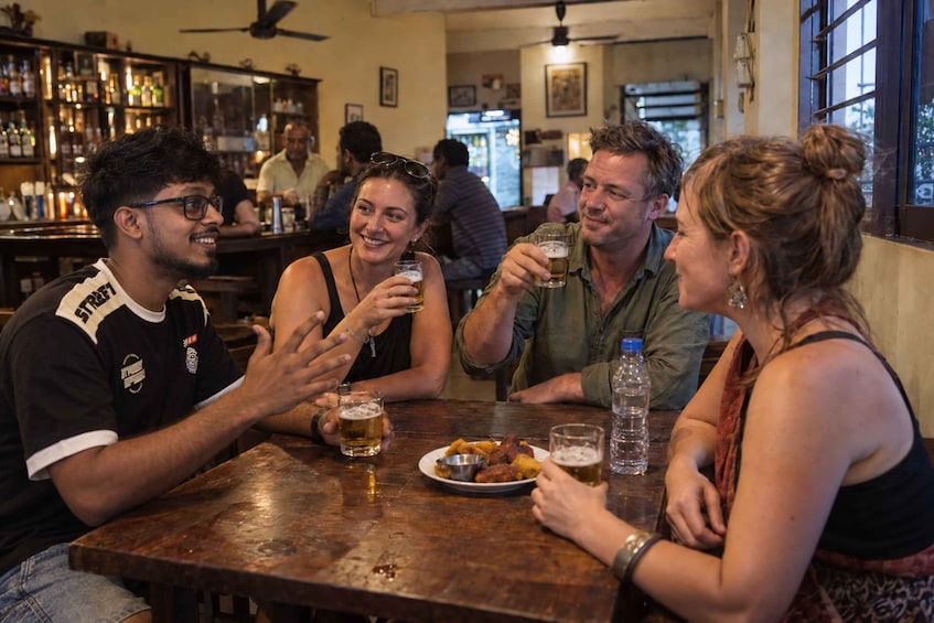 A group of people sitting at a wooden table in a bar with bottles and glasses. 1