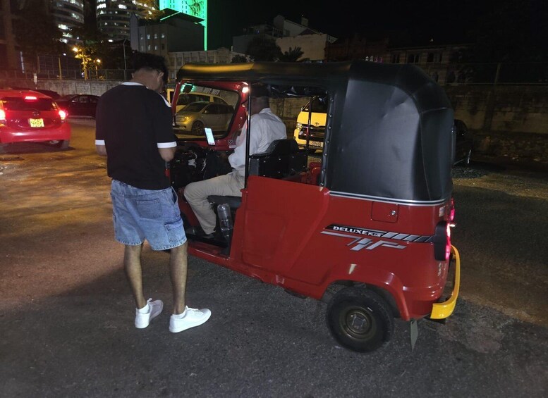 A red auto-rickshaw with a man standing beside it, another man inside, and a car in the background. 3