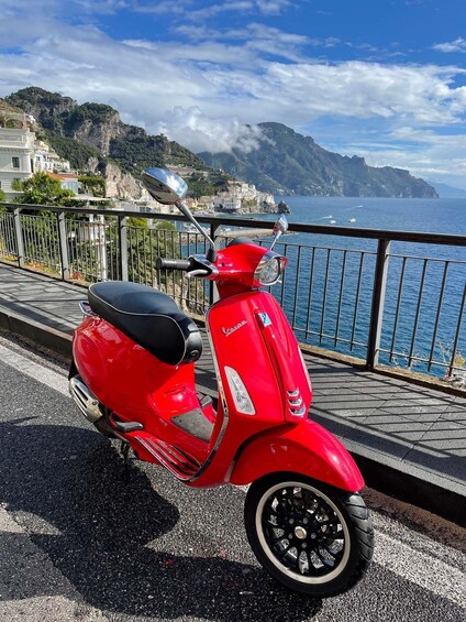 A red Vespa scooter parked on a paved area with a scenic coastal view in the background. 1
