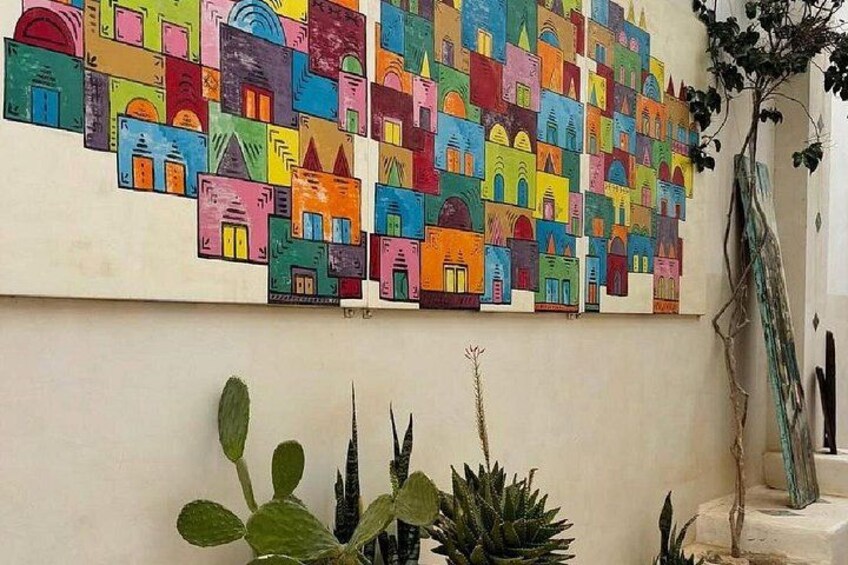 Una pintura abstracta y colorida de casas sobre una pared, rodeada de plantas en macetas. 5