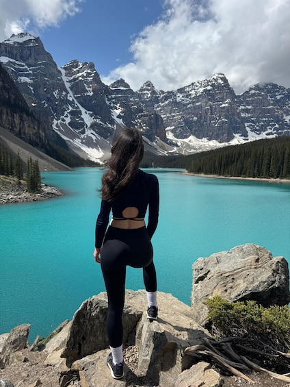 A person standing on a rocky outcrop overlooking a turquoise lake with snow-capped mountains in the background. 5