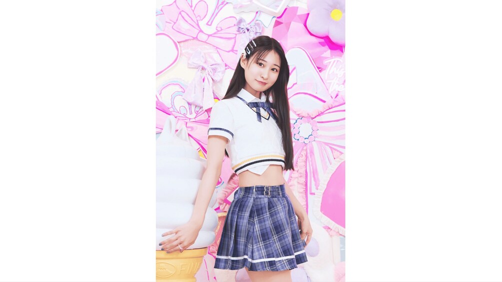 A person in a white and blue sailor-style top and plaid skirt, standing in front of a pink and white floral background. 5