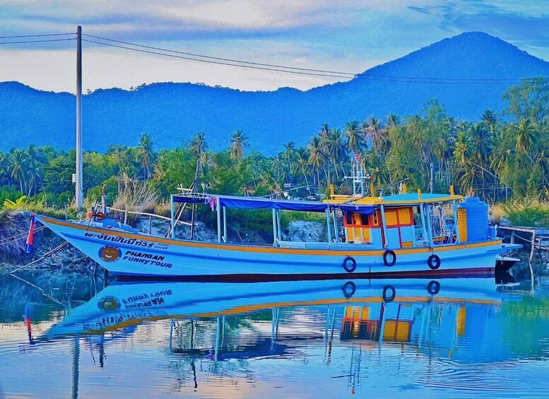 A blue and yellow boat named PHAMGAN FUNNY TOUR docked at a calm water body with palm trees and a mountain in the background. 1