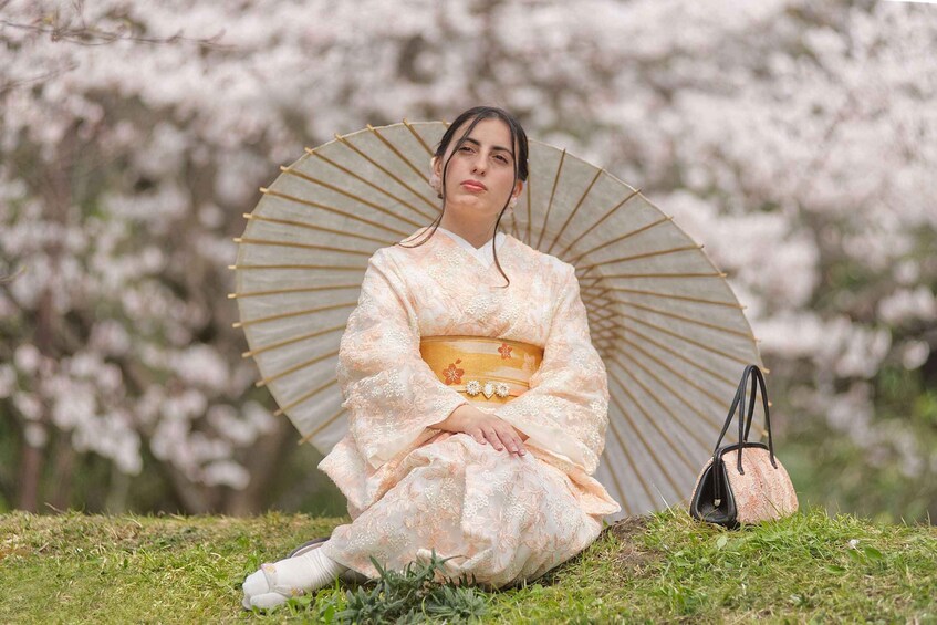 A person in traditional attire sitting on grass with a fan and a bag. 4