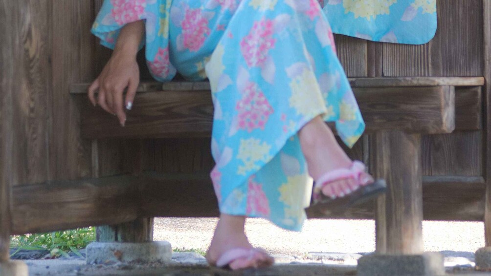 A person sitting on a wooden bench wearing a floral dress and sandals. 2