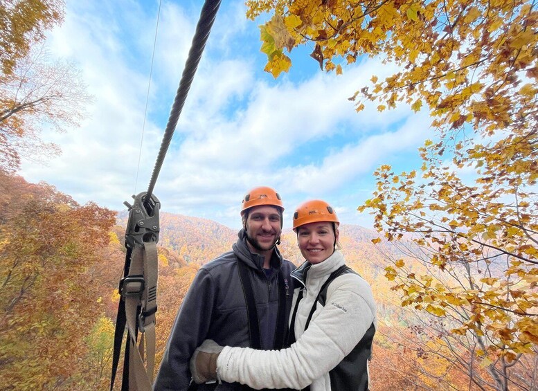 Two people wearing helmets and harnesses, standing on a zip line with autumn foliage in the background. 2