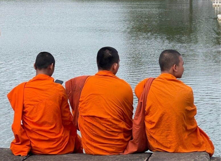 Three monks in orange robes sitting by a calm body of water. 4