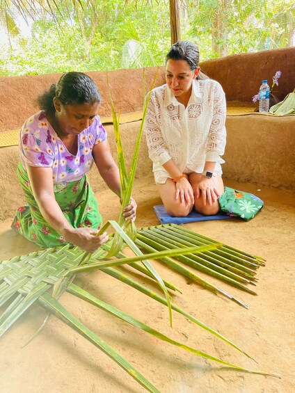 Two people engaged in weaving with green leaves on a sandy floor. 4