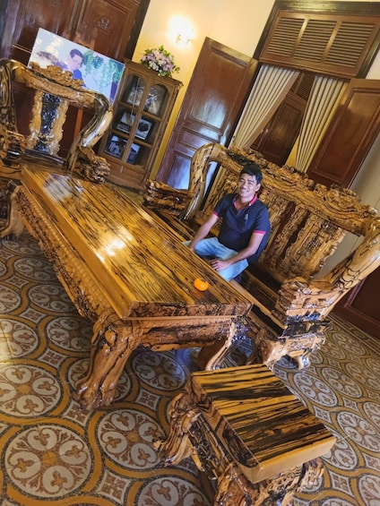 A man sitting on an ornate, intricately carved wooden sofa with a large, polished wooden coffee table in front of him. 2