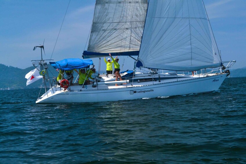 A sailboat with a crew on a calm sea, a flag, and a mountainous landscape in the background. 1