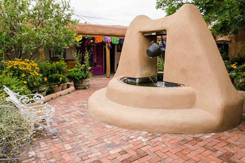 A rustic outdoor area with a stone fountain, a white metal bench, and a building with colorful awnings. 2